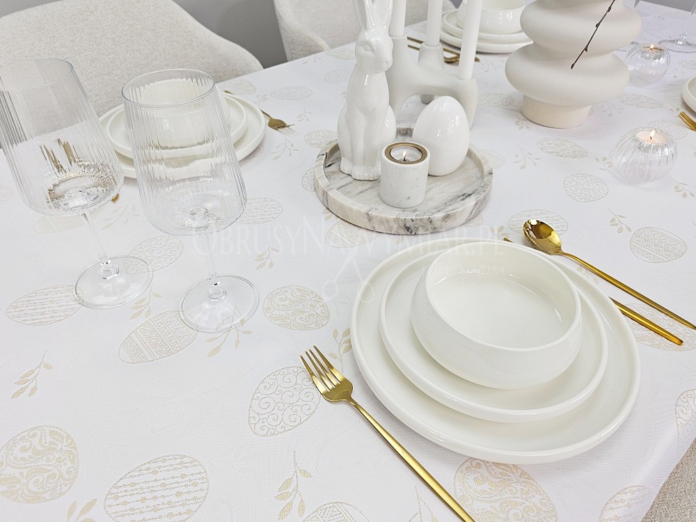 Aranżacja stołu wielkanocnego biały obrus plamoodporny ze złotymi pisankami, biała zastawa i złote sztućce